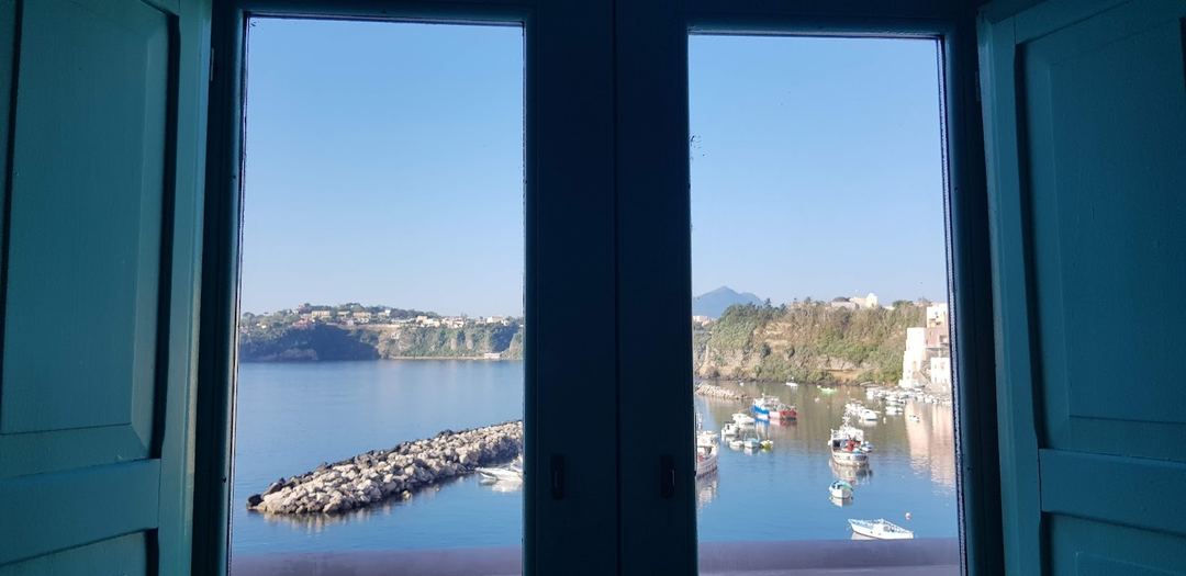 A scenic view of a coastal harbor with moored fishing boats, rocky breakwater, and tree-lined cliffs framed through turquoise-blue window shutters.