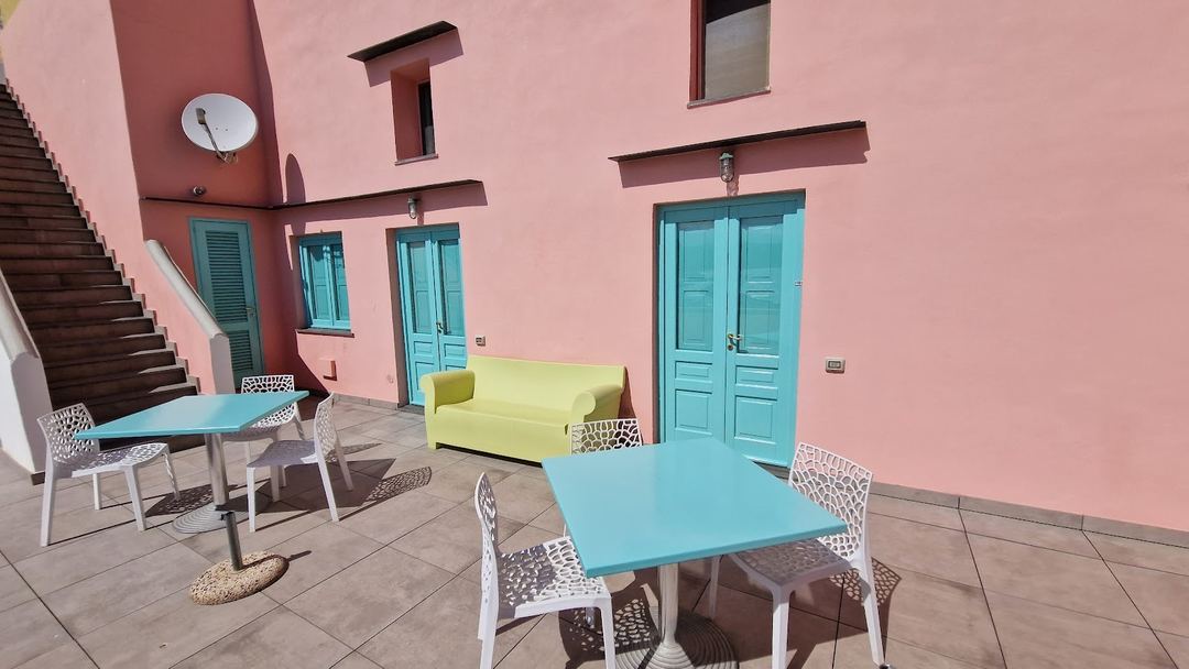 A bright courtyard with colorful seating areas featuring turquoise tables, white patterned chairs, and a lime green sofa against a pink building facade.